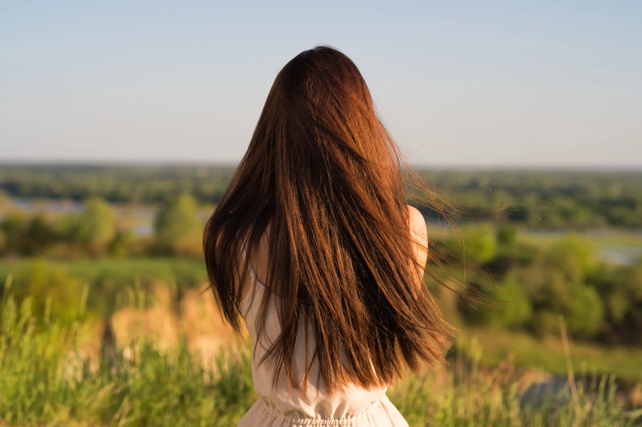 Young girl sits with his back to the camera and looks at nature. Peaceful girl siting  by a cliff enjoying the landscape. - outdoors. Freedom concept. Long brown hair. Rear view